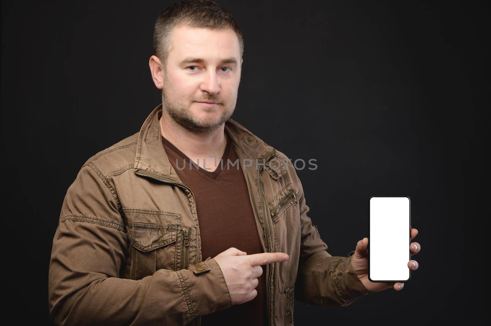 Studio portrait of a casual plump man in a yellow sweater and cap holding an smartphone with a white cut-out screen in his hands. Website presentation or application copy space.