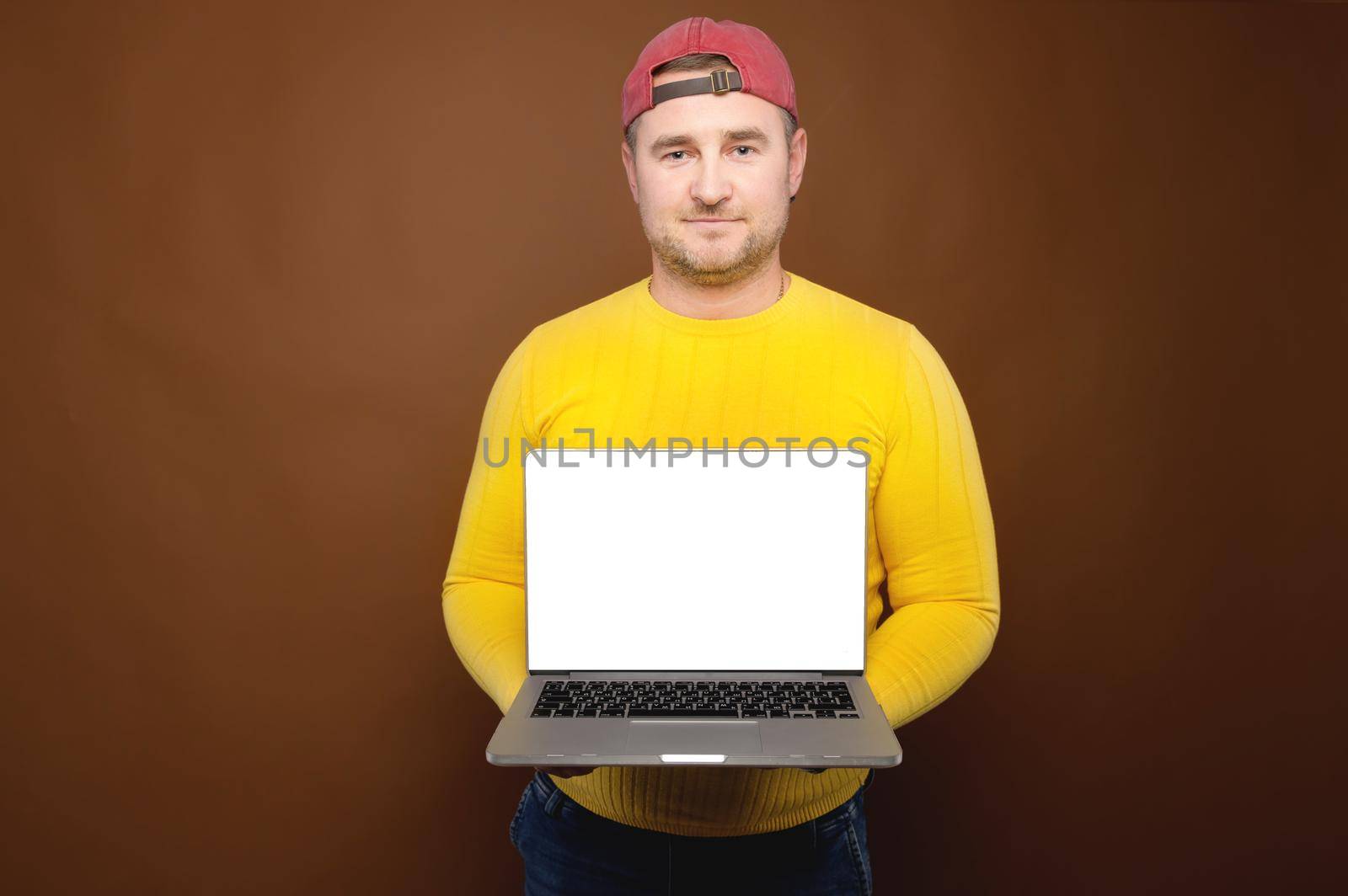 Studio portrait of a casual plump man in a yellow sweater and cap holding an open laptop with a white cut-out screen in his hands. Website presentation or application copy space.