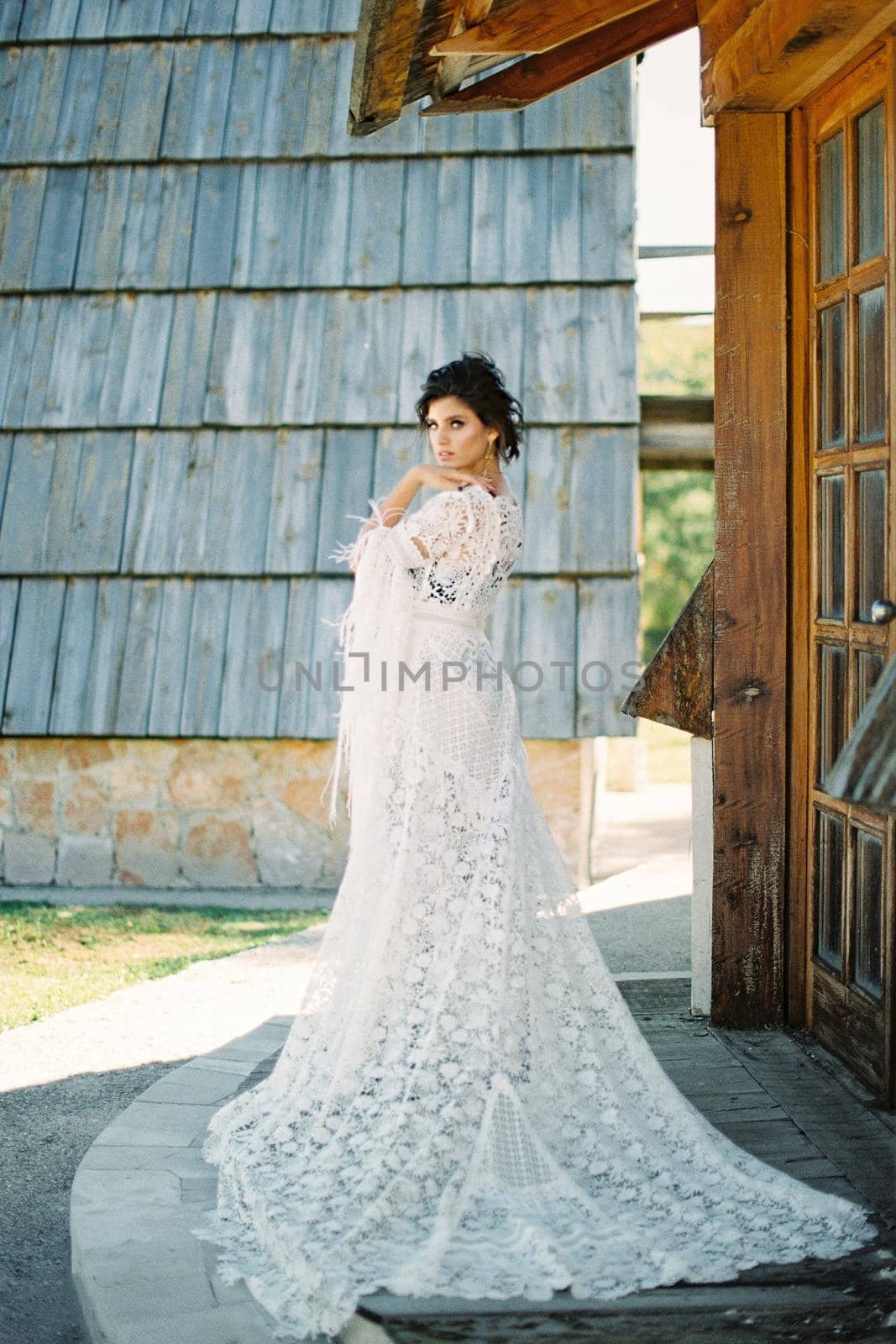 Bride in a long white wicker dress stands on the threshold of a wooden house. High quality photo