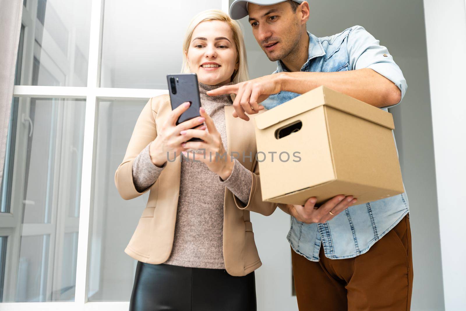 Fast and reliable service. Cheerful young delivery man giving a cardboard box to young woman while standing at the entrance of her apartment by Andelov13