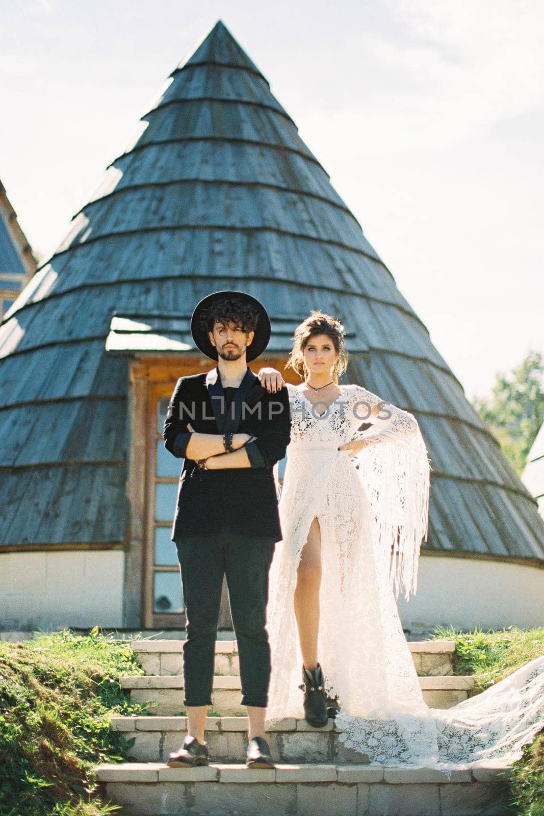 Bride put her hand on the shoulder of groom standing on the steps near the conical house. High quality photo