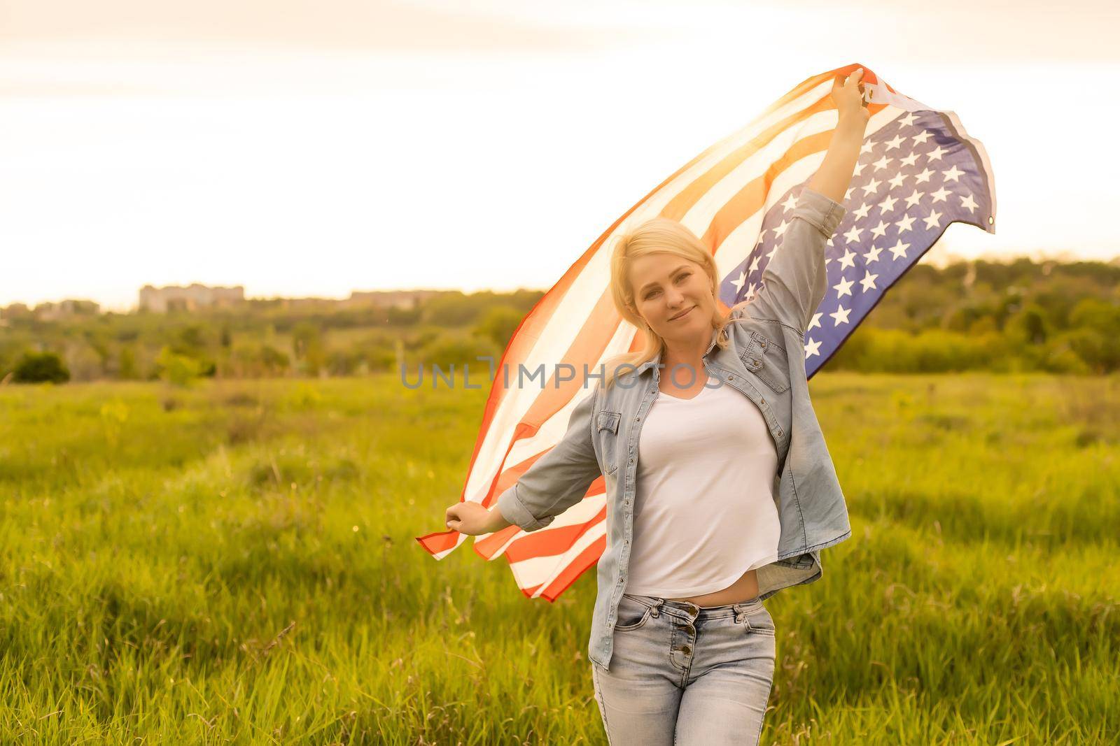 attractive woman holding an American flag in the wind in a field. Summer landscape against the blue sky. Horizontal orientation