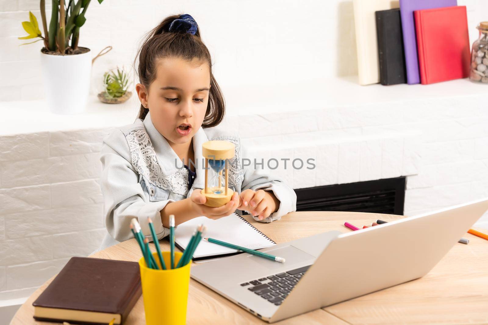 little girl with a laptop at the table and holds an hourglass, waiting for the end of online lessons