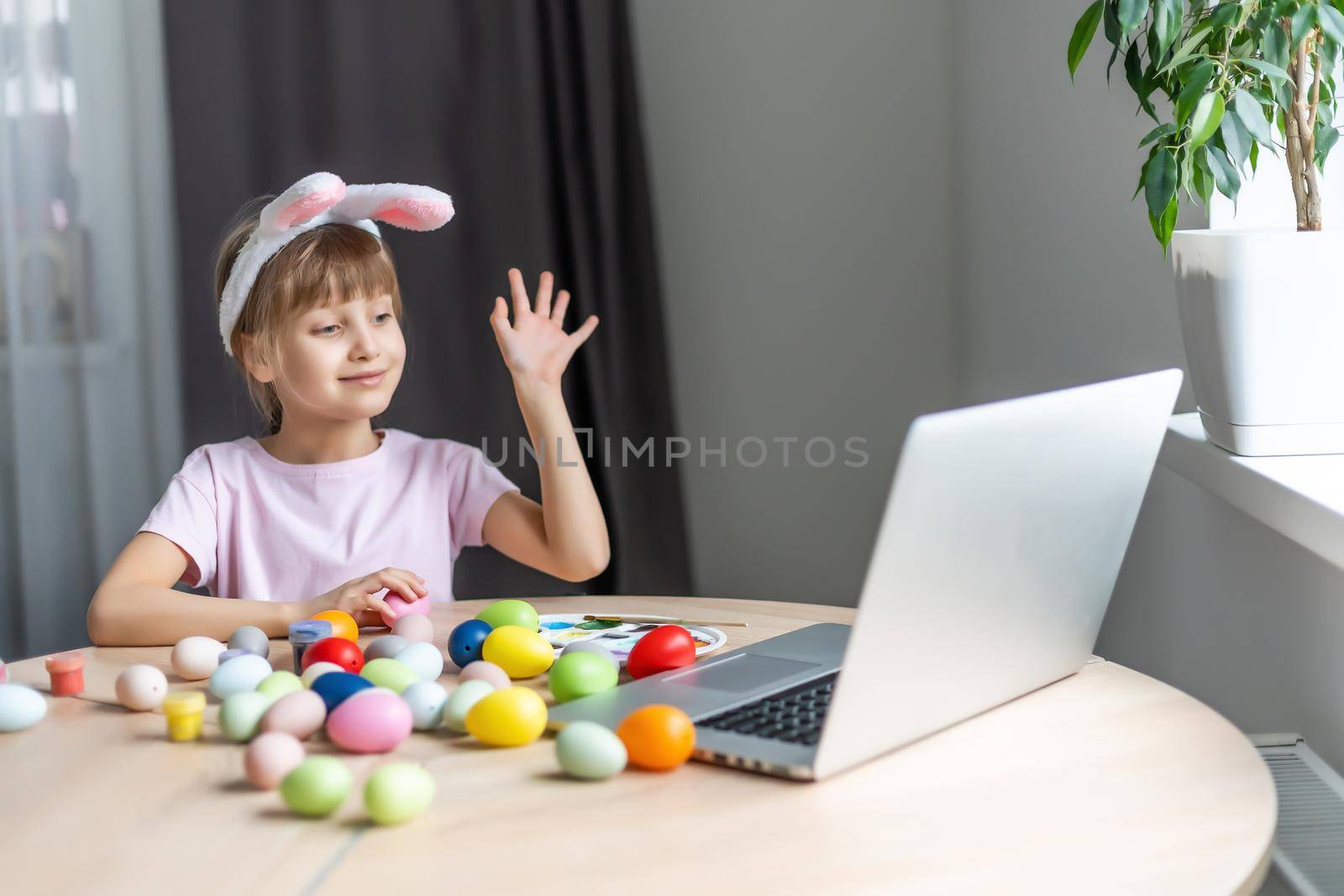 Pretty little girl wearing bunny ears and painting easter eggs at home during coronavirus covid-19 outbreak. Kid using laptop on kitchen, online, video call to friends.