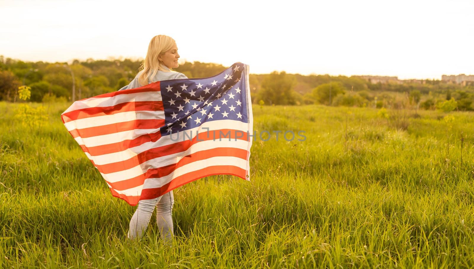 attractive woman holding an American flag in the wind in a field. Summer landscape against the blue sky. Horizontal orientation