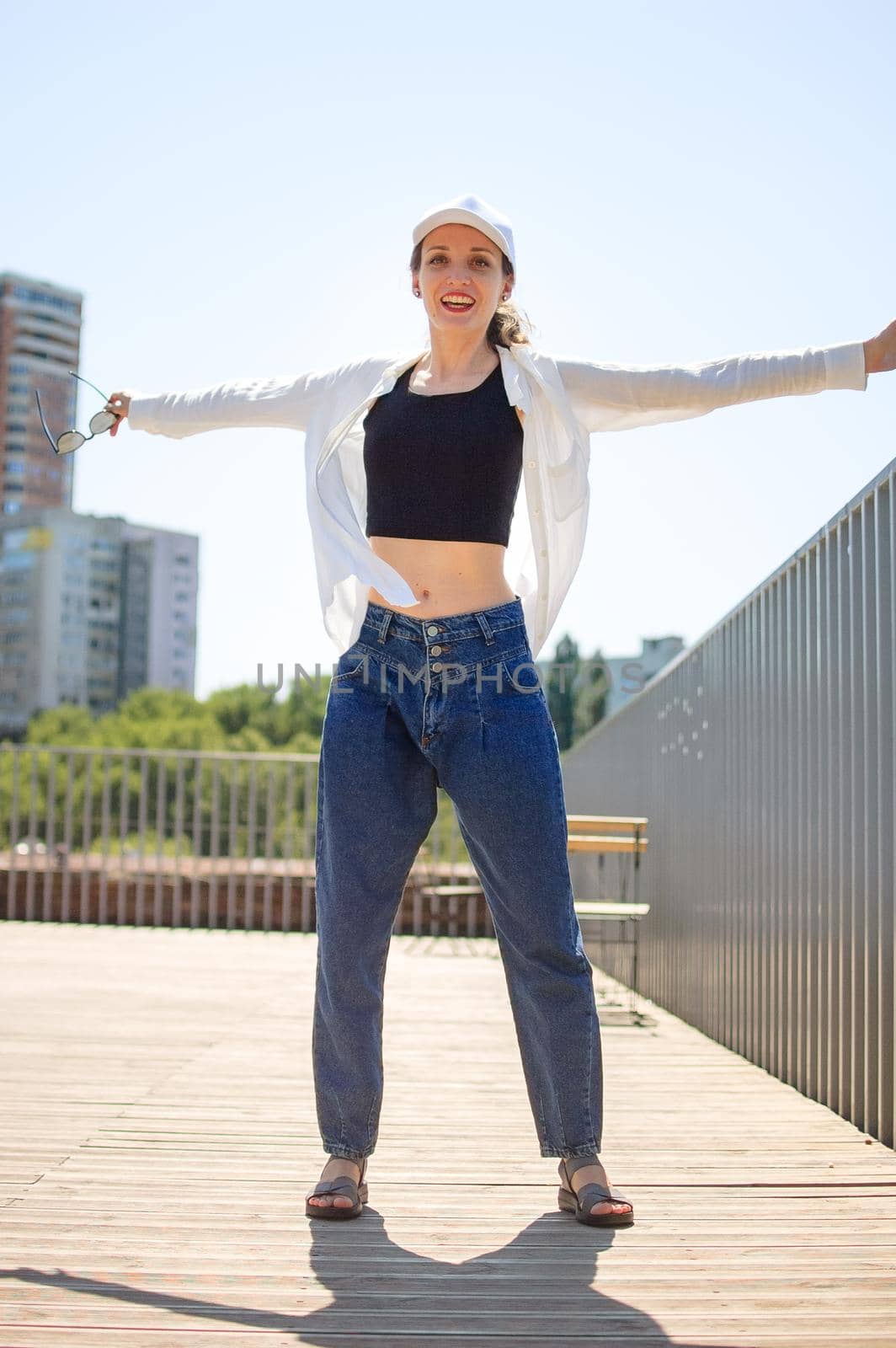 Female portrait of young active girl in black top, white shirt, basketball cap, and jeans on modern buildings background outdoors by balinska_lv