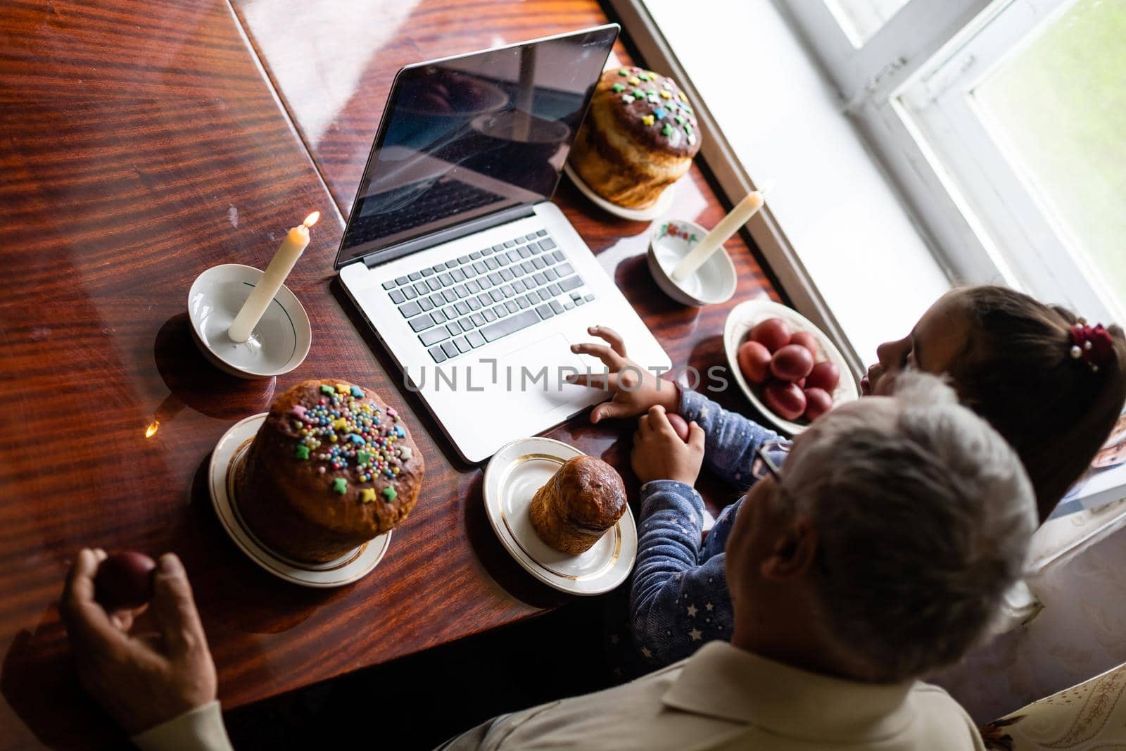 grandfather and granddaughter are talking via video link to their friends. Decorated table with colorful eggs and cake. Chatting during the COVID pandemic and the Easter holidays.