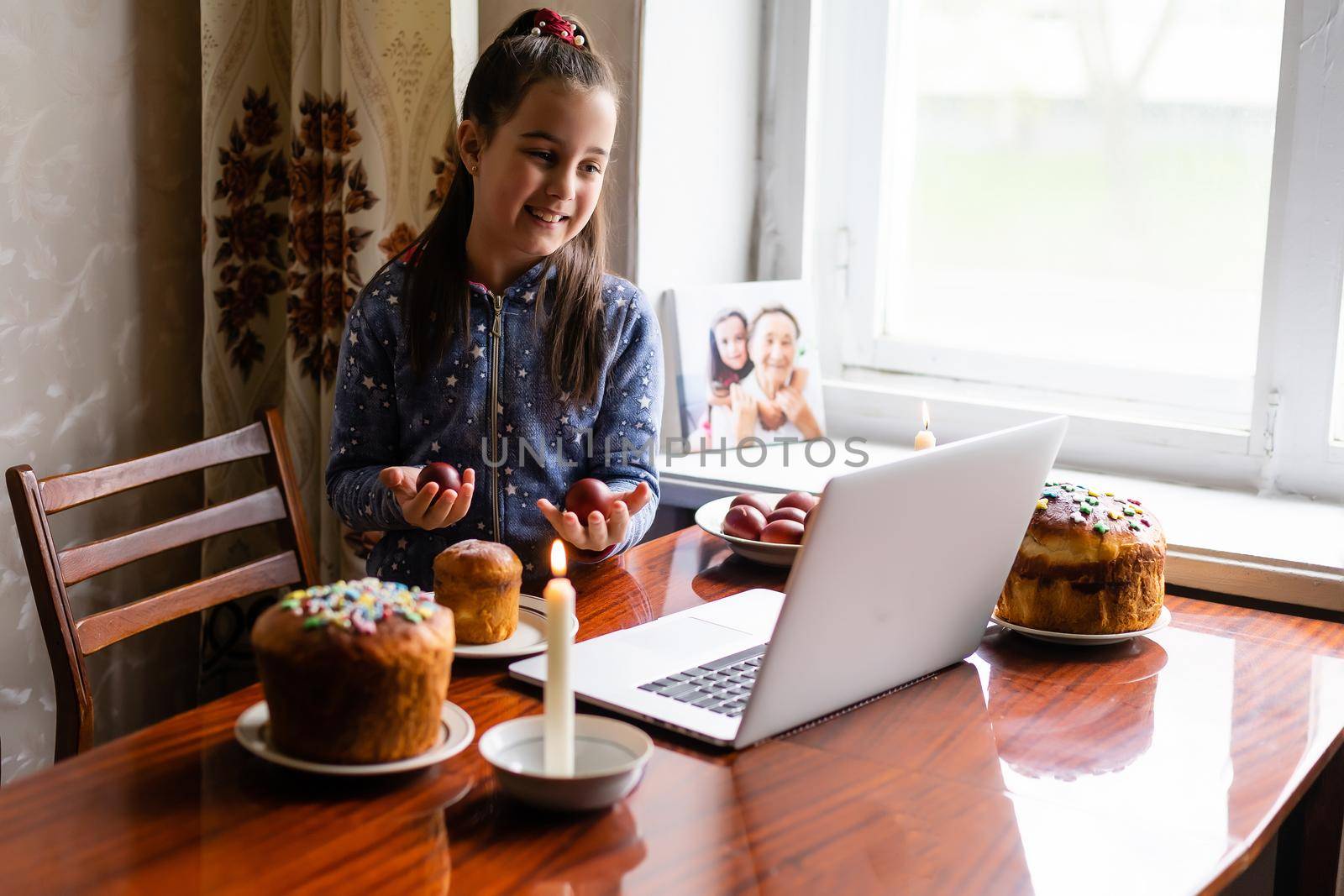 Pretty little girl painting easter eggs at home during coronavirus covid-19 outbreak. Kid using laptop on kitchen, online, video call to friends by Andelov13