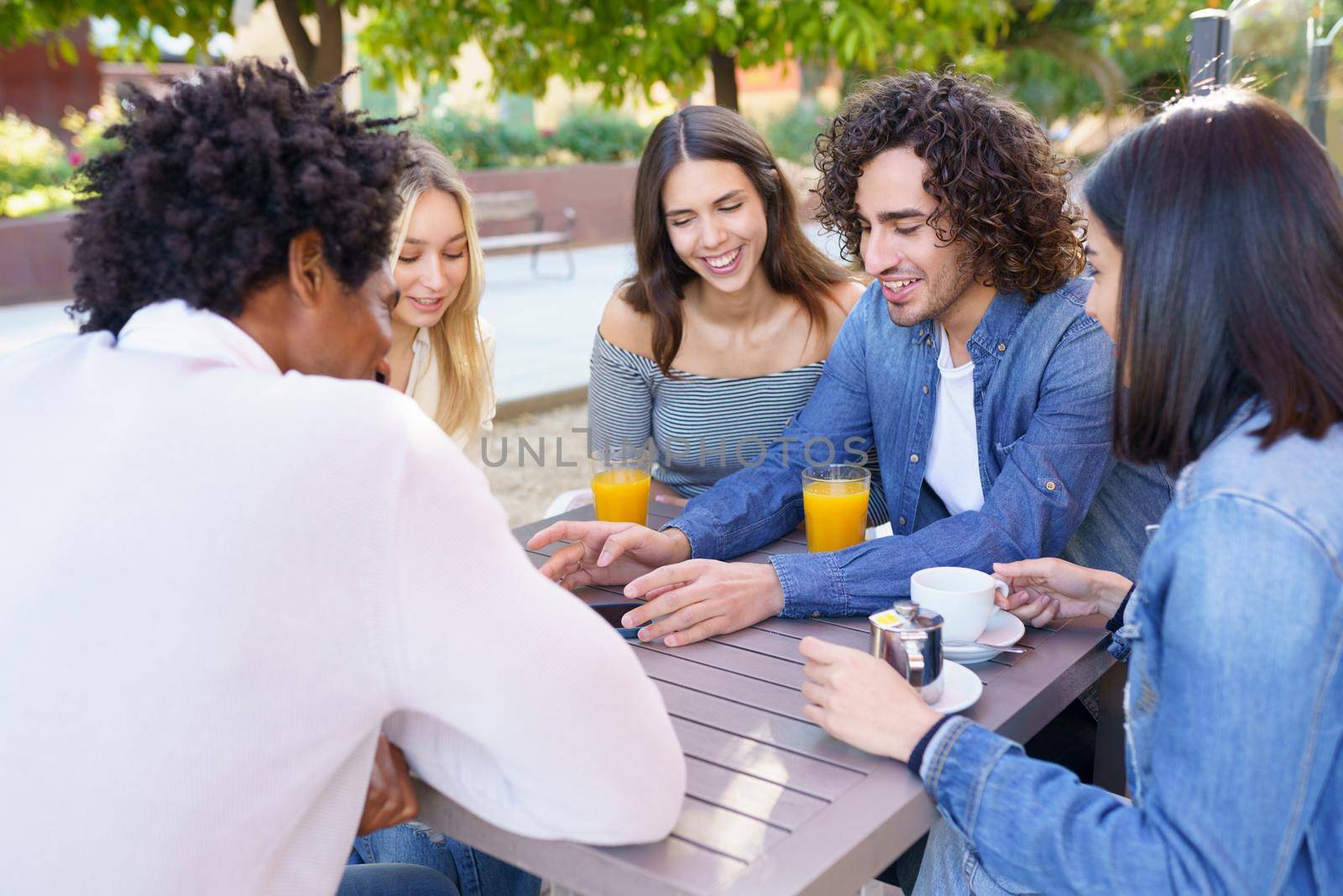 Company of happy multiethnic friends gathering at wooden table in summer cafe and spending time together at weekend