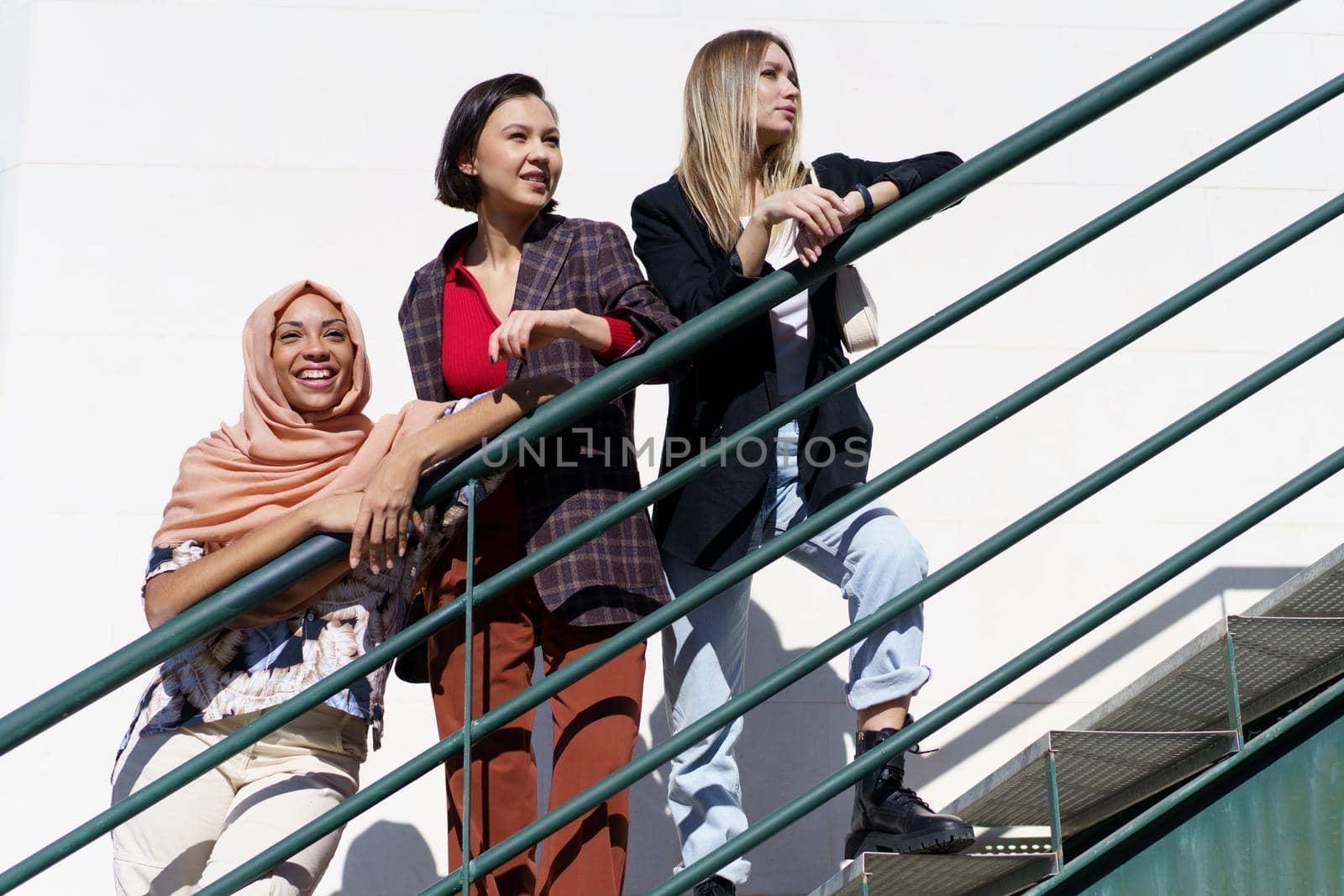 Low angle of trendy young multiethnic female best friends, on stylish outfit and hijab, leaning on metal railing while standing on staircase and looking away in city on sunny day