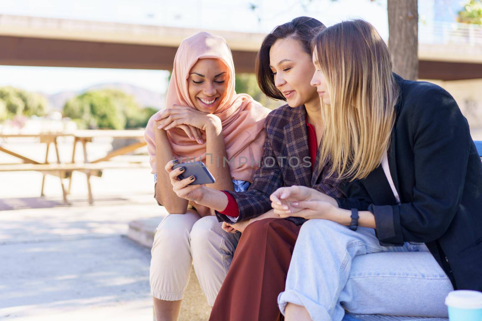 Positive multiethnic girlfriends sharing smartphone while resting on bench in park by javiindy