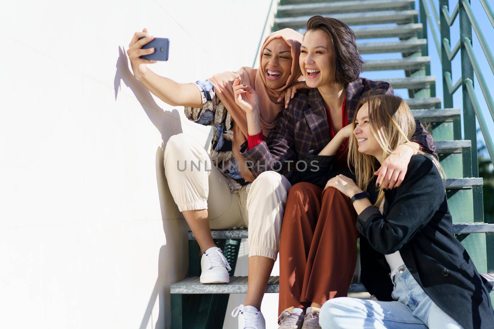 Trendy young cheerful Muslim lady in stylish outfit and traditional hijab, smiling brightly while taking selfie on mobile phone, sitting on staircase with diverse girlfriends on sunny day