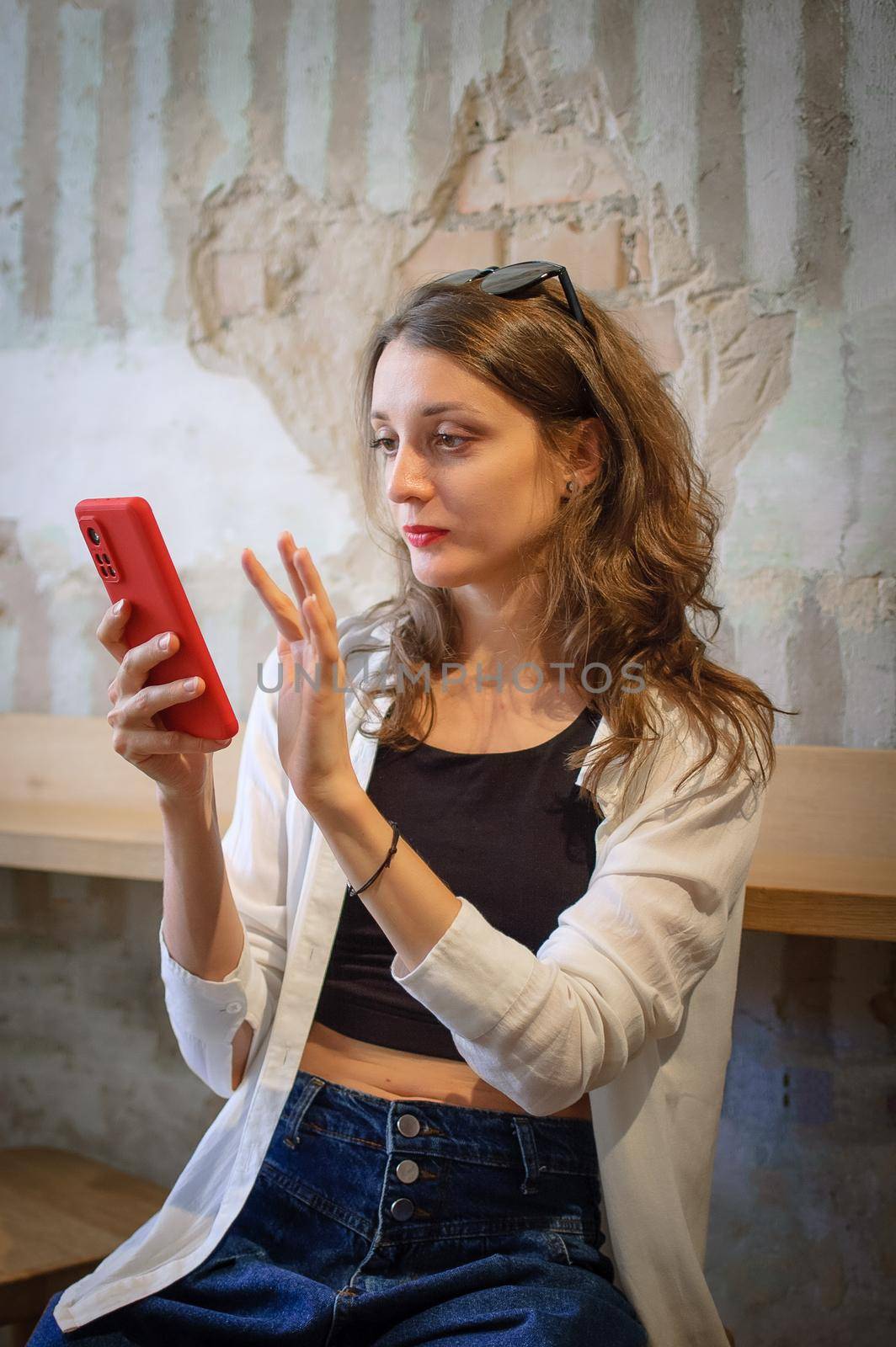 Young woman in white shirt and black top is browsing using her red smartphone to make online shopping or payment sitting indoors on loft wall background, technology, happy people concept by balinska_lv