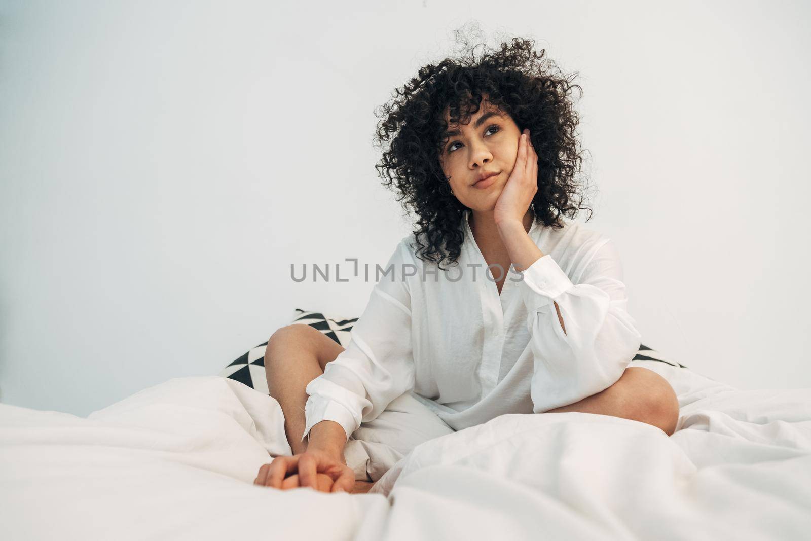 Pensive young mixed race woman with curly hair sitting on bed resting face on hand. Copy space. Lifestyle concept.