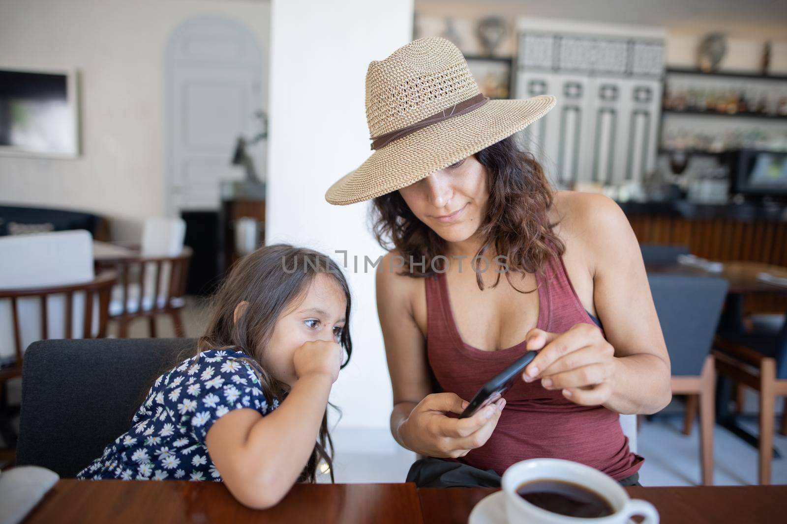 Portrait of beautiful mother with hat showing smartphone to adorable little girl in restaurant. Brunette woman and curious daughter watching modern mobile phone in cafe. Family and technology