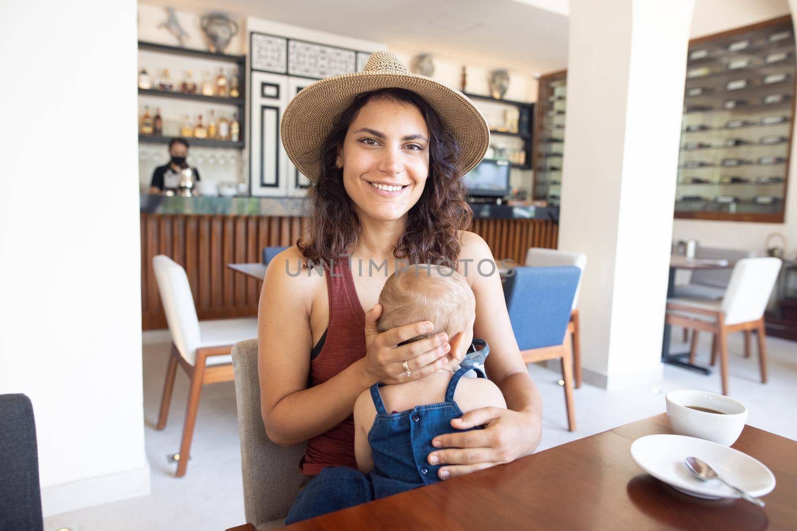 Portrait of beautiful smiling mother wearing a hat and holding adorable baby in restaurant. Happy brunette woman and young daughter at table with cafe counter as background. Lovely family on holiday