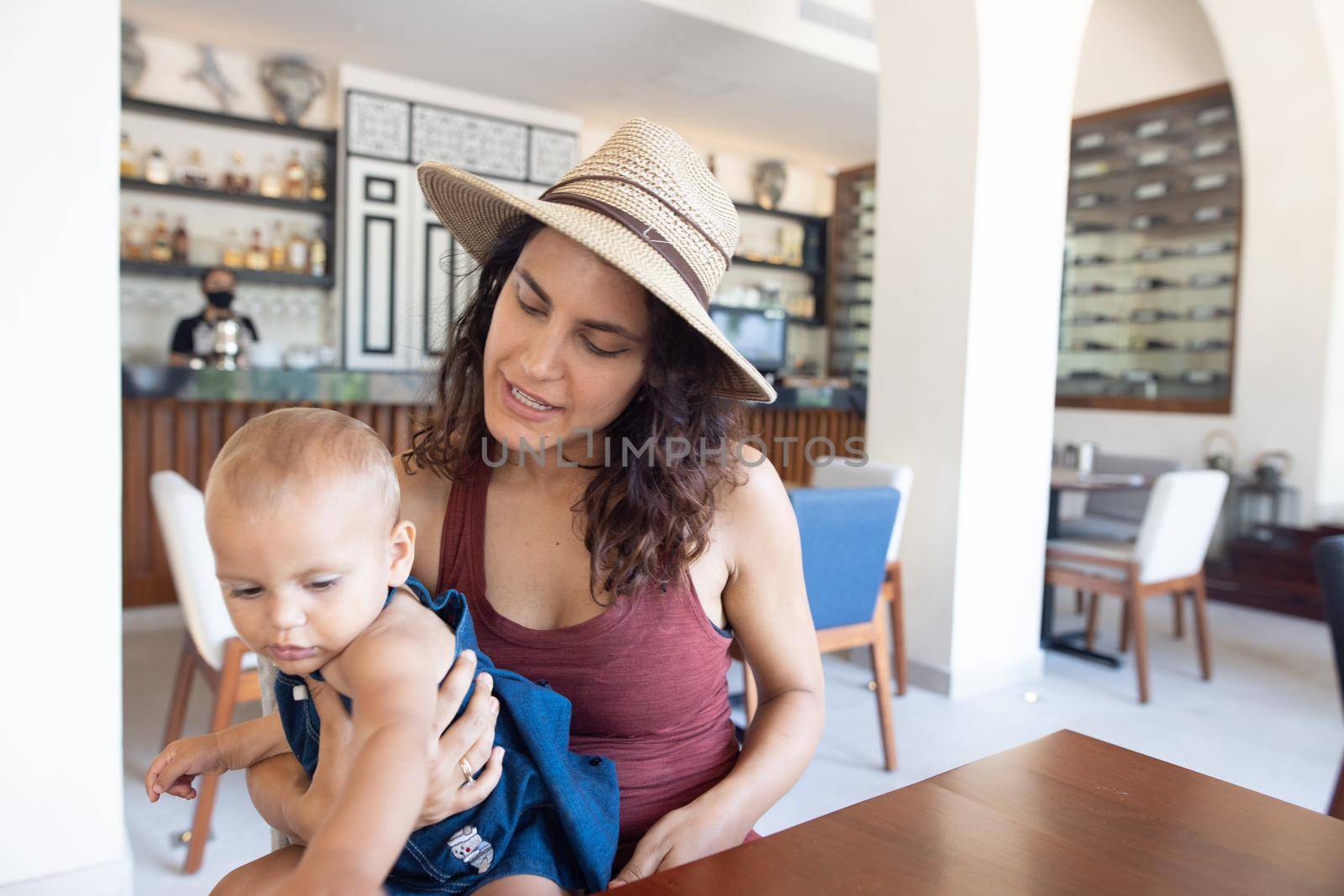 Portrait of beautiful smiling mother wearing a hat and holding adorable baby in restaurant. Happy brunette woman and young daughter at table with cafe counter as background. Lovely family on holiday