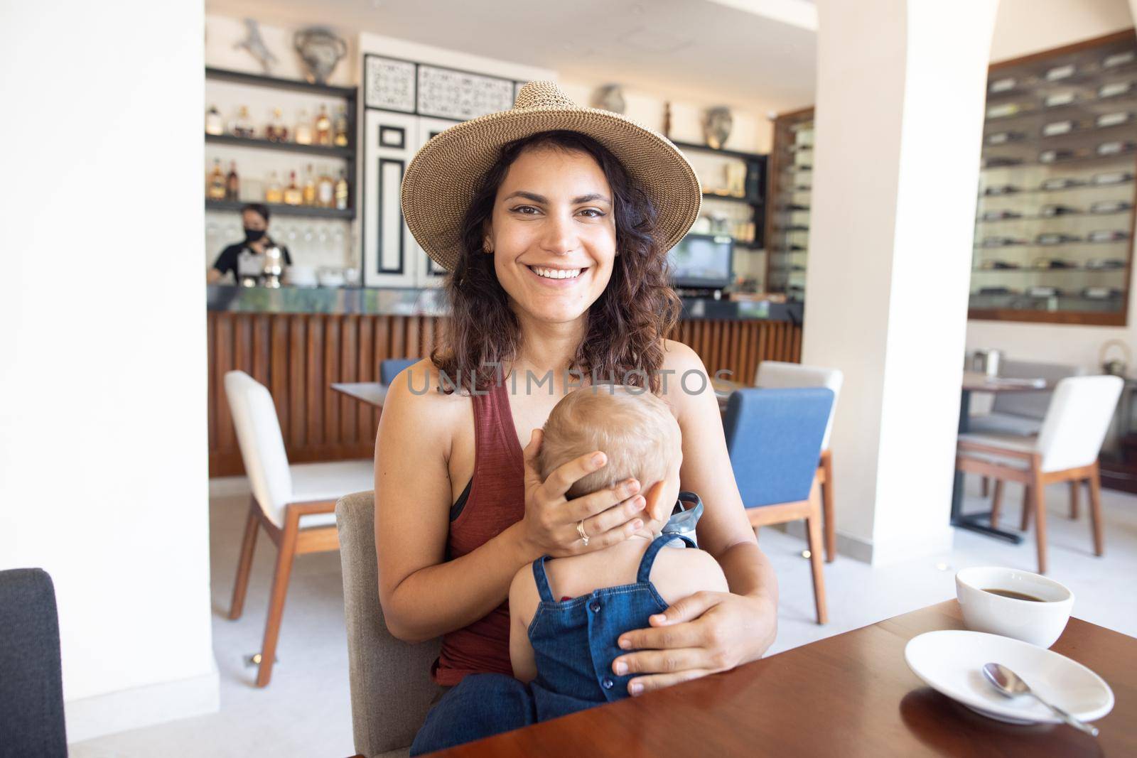 Portrait of beautiful smiling mother wearing a hat and holding adorable baby in restaurant. Happy brunette woman and young daughter at table with cafe counter as background. Family having breakfast