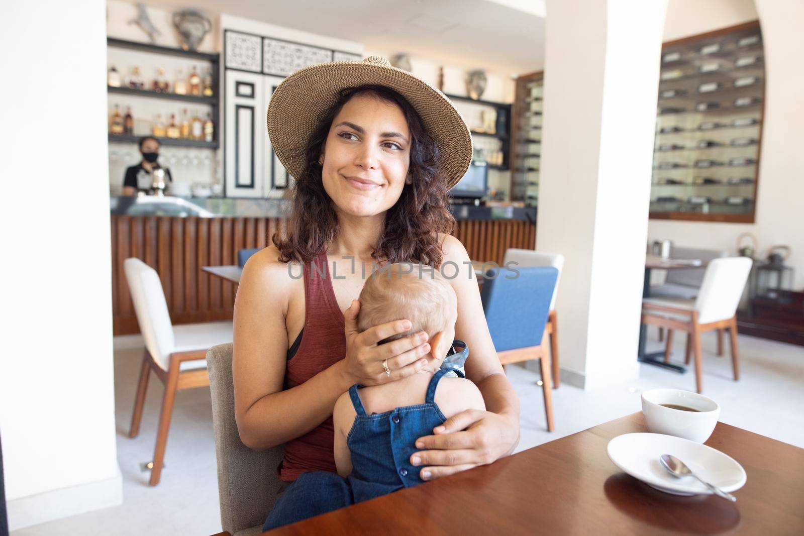 Portrait of beautiful smiling mother wearing a hat and holding adorable baby in restaurant. Happy brunette woman and young daughter at table with cafe counter as background. Lovely family on holiday