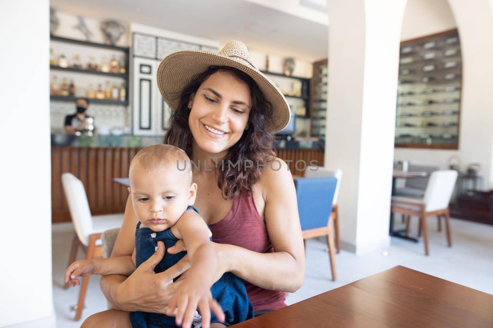 Portrait of beautiful smiling mother wearing a hat and holding adorable baby in restaurant. Happy brunette woman and young daughter at table with cafe counter as background. Family having breakfast