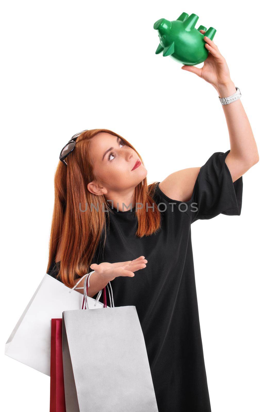 Young redhead girl holding shopping bags and an empty piggy bank, with surprised expression on her face, isolated on white background. Overspending concept. Financial debt concept.