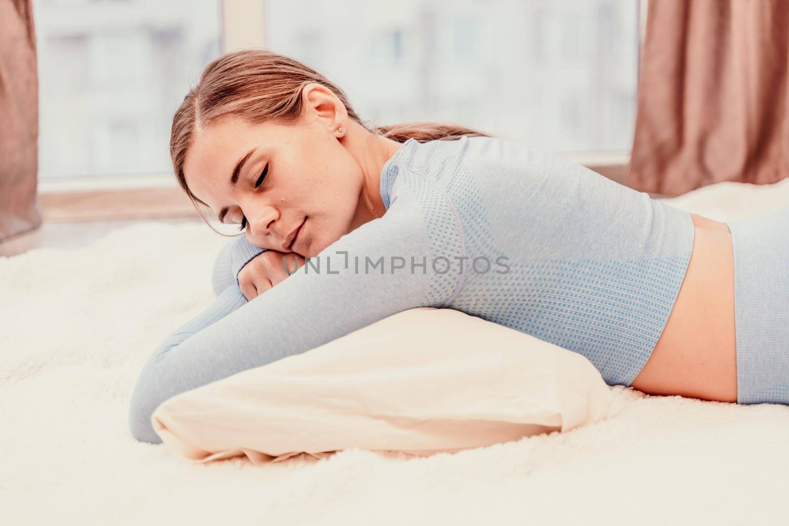 Side view portrait of relaxed woman with long hair lying on carpet at home. She is dressed in a blue tracksuit, holding a phone in her hands