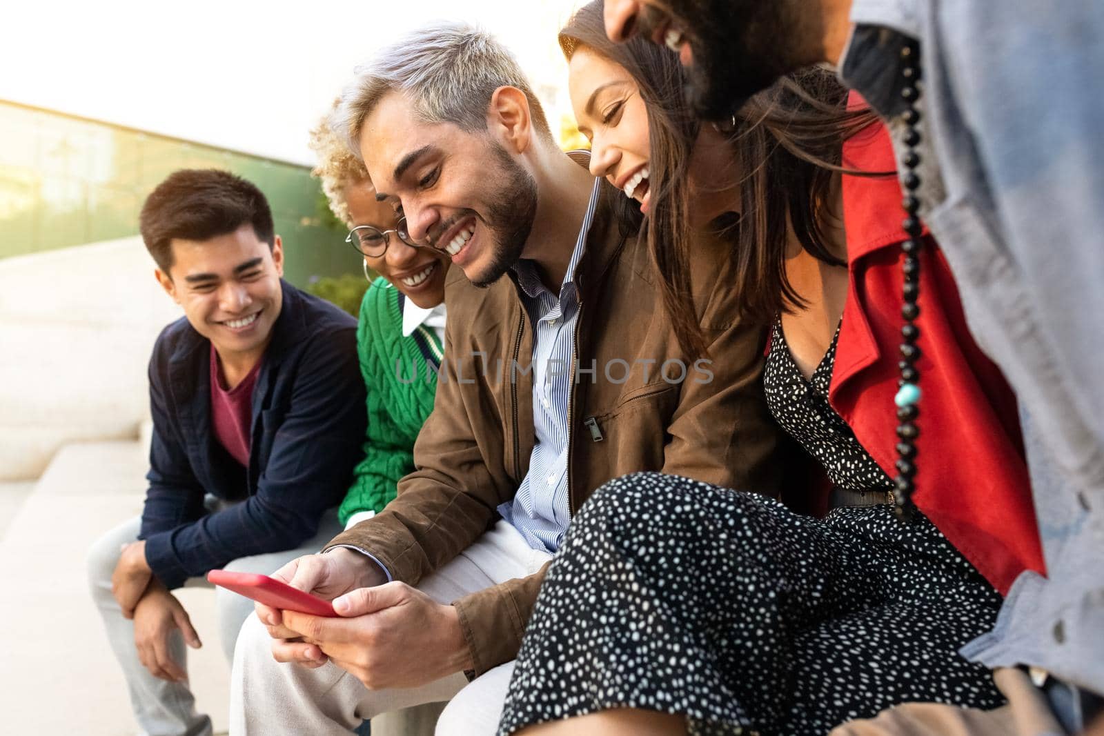 Group of millennial multiracial friends sit on a bench looking at cellphone together. Watching online video. by Hoverstock