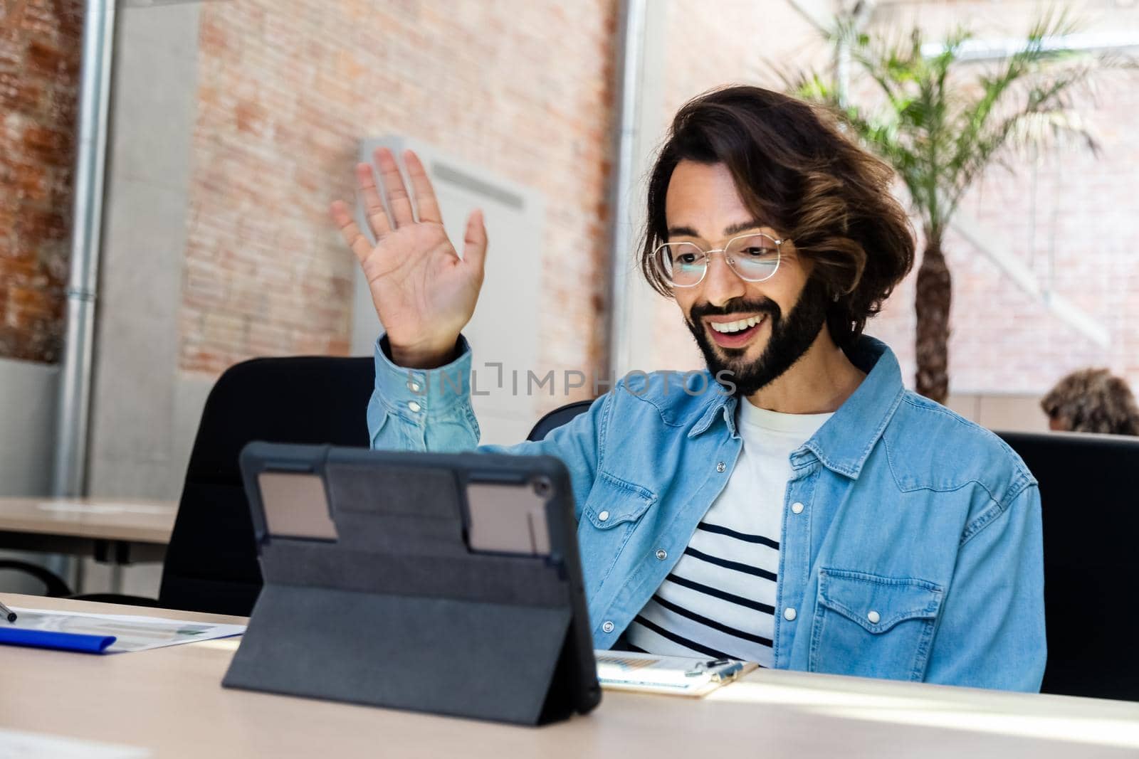 Smiling young caucasian man waving hello on a video call using a digital tablet in the office. Copy space. Technology concept.