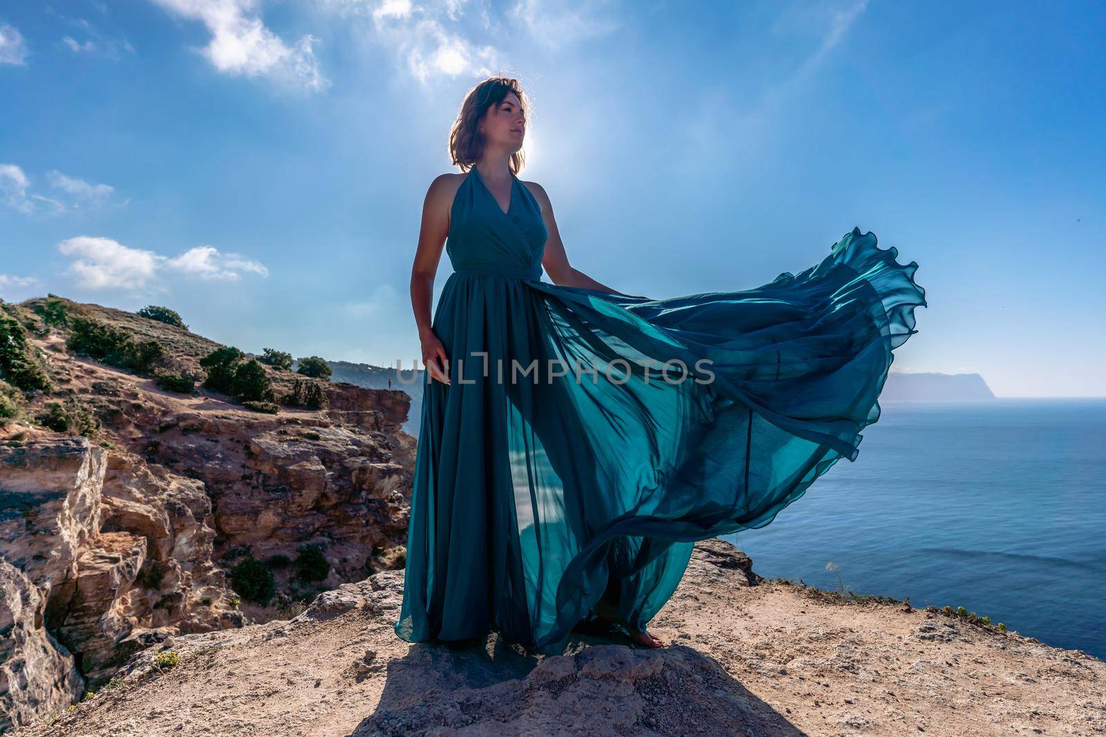 A girl with loose hair in a long mint dress descends the stairs between the yellow rocks overlooking the sea. A rock can be seen in the sea. Sunny path on the sea from the rising sun.