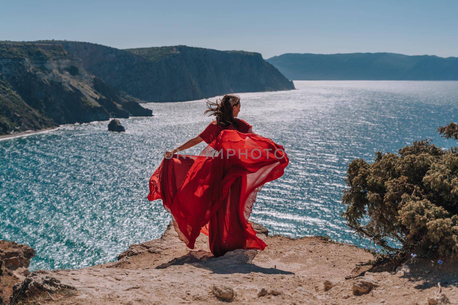 A girl with loose hair in a red dress waved her skirt on the yellow rocks overlooking the sea. Sunny path on the sea from the rising sun. by Matiunina