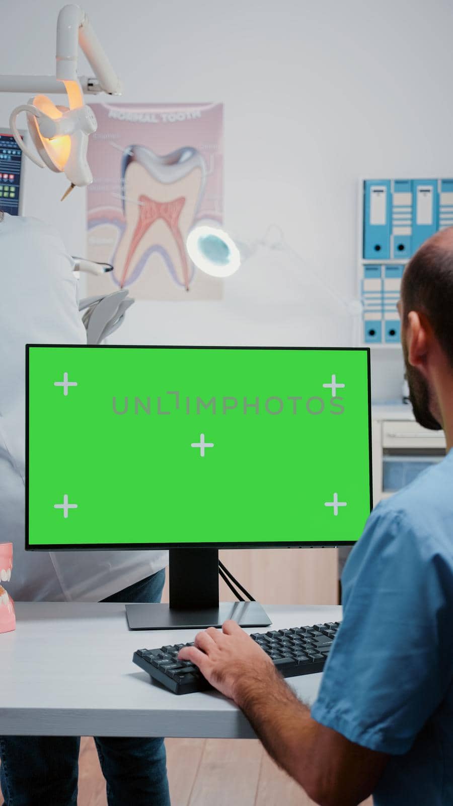 Vertical video: Man nurse working with keyboard and monitor with green screen. Dentistry assistant looking at chroma key with mockup template on monitor for stomatological care.