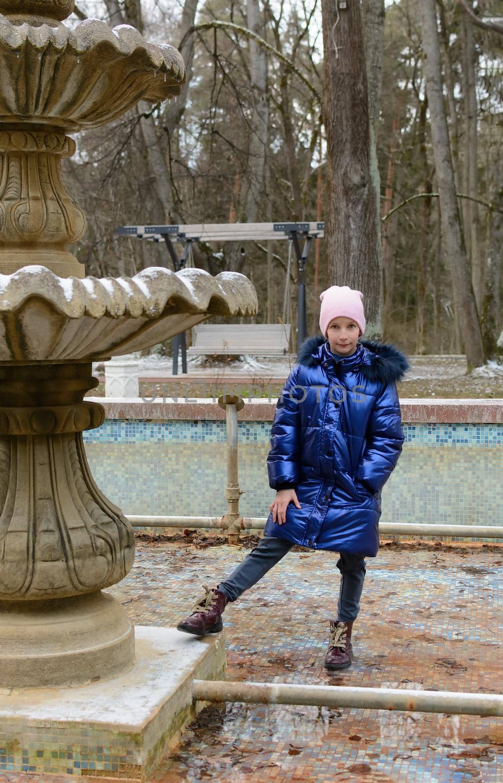 Girl in a blue coat stands in the dry bowl of a fountain in the park