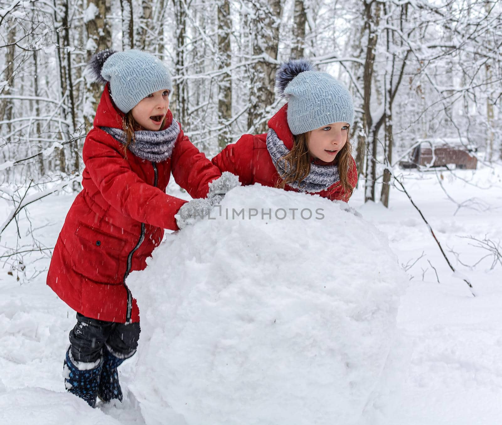 Two twin girls in red jackets rolling huge snowball to make snowman by andrew_donnikov