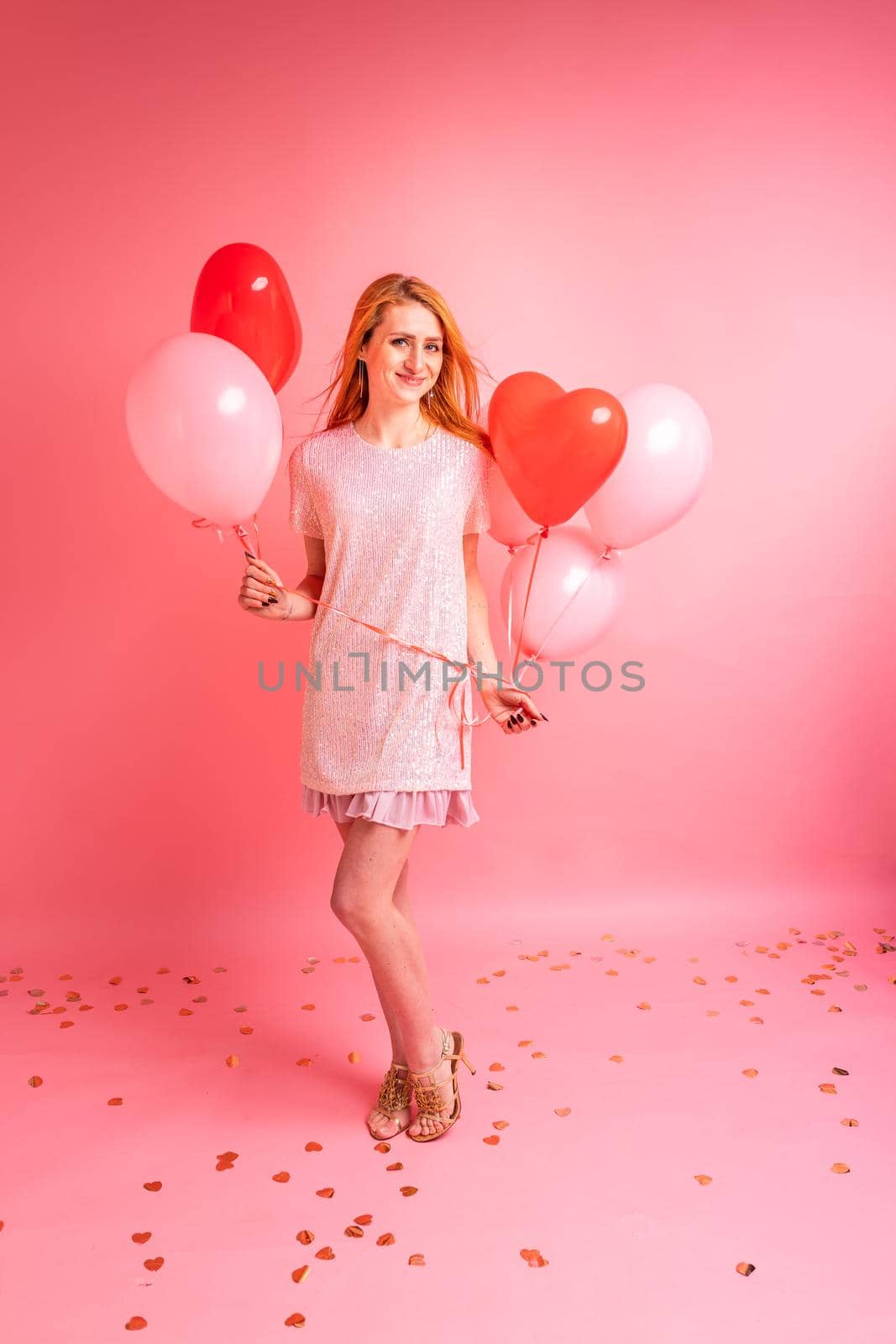 Beautiful redhead girl with red heart baloon posing. Happy Valentine's Day concept. Studio photo of beautiful ginger girl dancing on pink background.