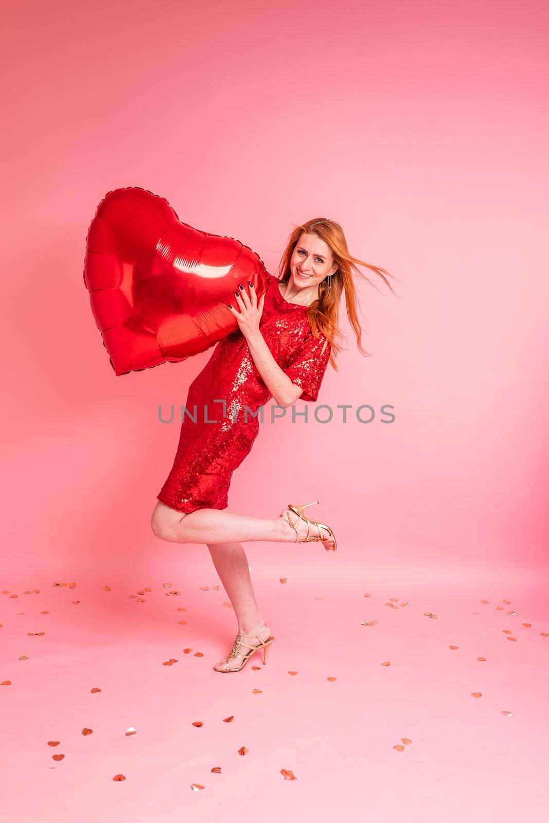 Beautiful redhead girl with red heart baloon posing. Happy Valentine's Day concept. Studio photo of beautiful ginger girl dancing on pink background.