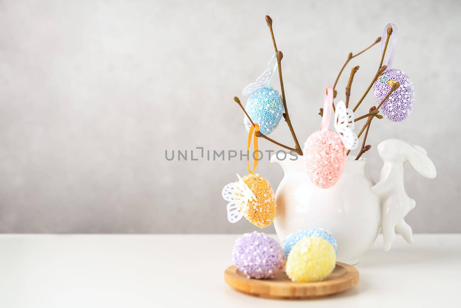 Home interior with easter decor. Vase with willow tree branches with Easter eggs and bunny on white table and background with copy space.