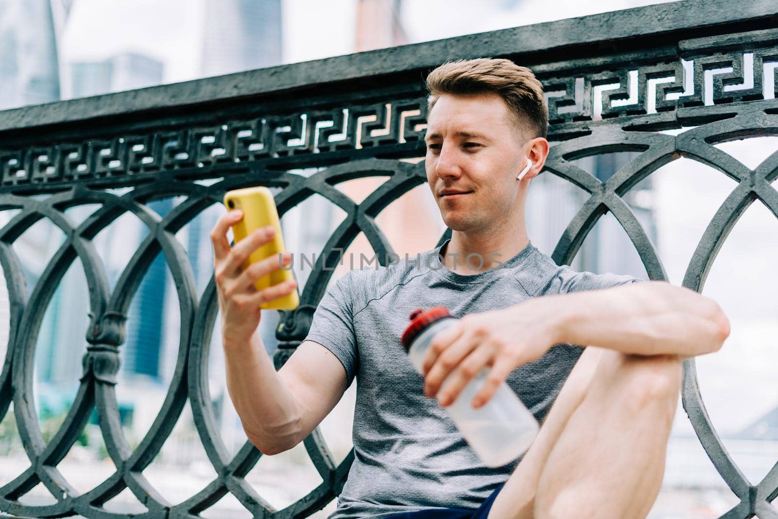 Tired Young man runner sitting on road, using mobile phone and relaxing after sport training. Holding water bottle while doing fitness workout in city urban street, cloudy sky at summer.