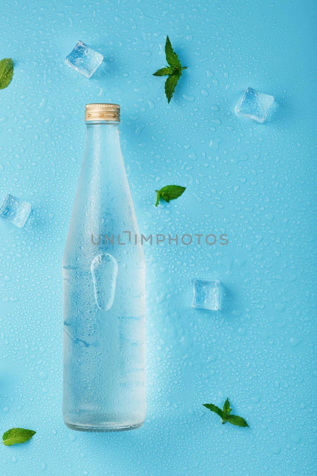 A bottle of ice water, ice cubes, drops and mint leaves on a blue background. Closed glass flask with a drink, layout. Free space, The view from the top