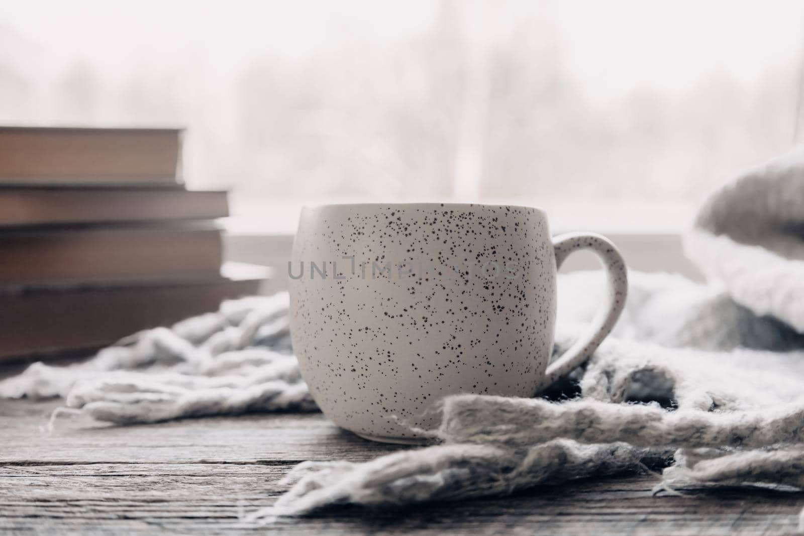 Minimalist style. Coffee, cozy grey sweater and books on vintage windowsill against snow landscape from outside. Soft focus. Relaxing winter day at home with traditional winter hot drink
