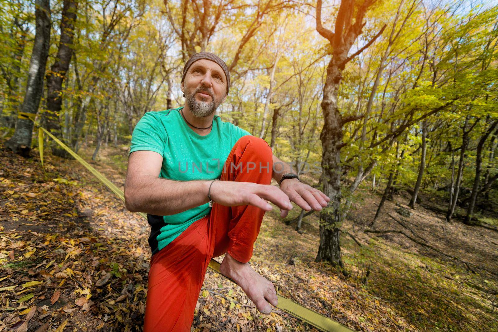 Portrait of a mature man in a scarf on his head sits on a taut slackline explains how to balance in the autumn forest.