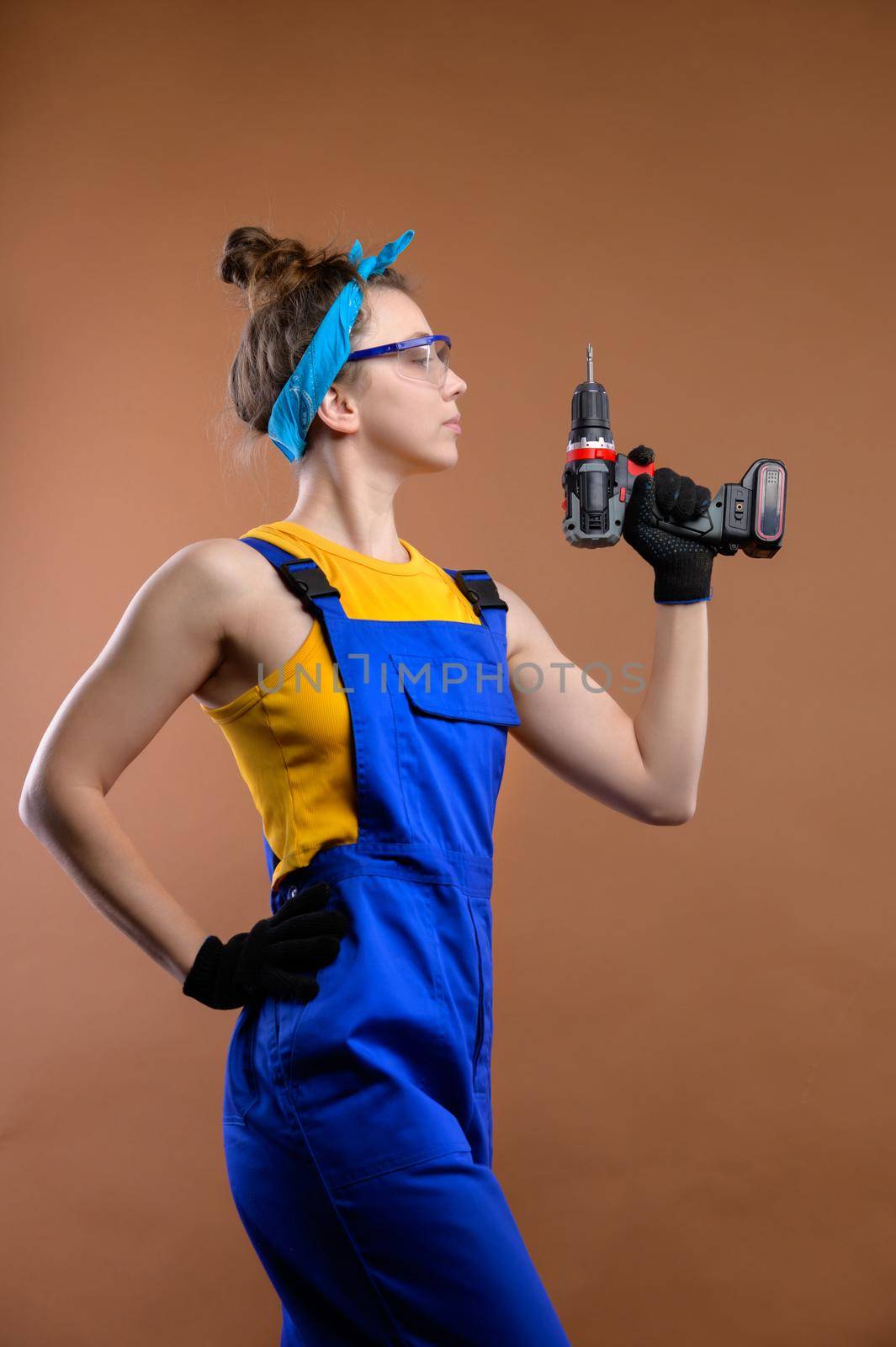 Young woman repairman in overalls and goggles works with a screwdriver in the studio on a brown background. Focus on the screwdriver. Copy space.