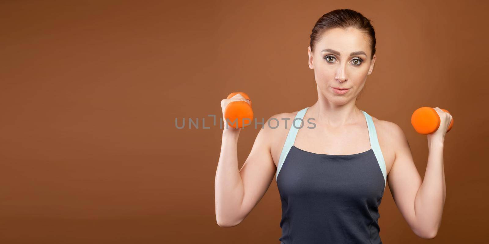 Panoramic shot of an athletic caucasian woman with dumbbells in her hands. Stands on a brown background with dumbbells in his hands and looks at the camera.