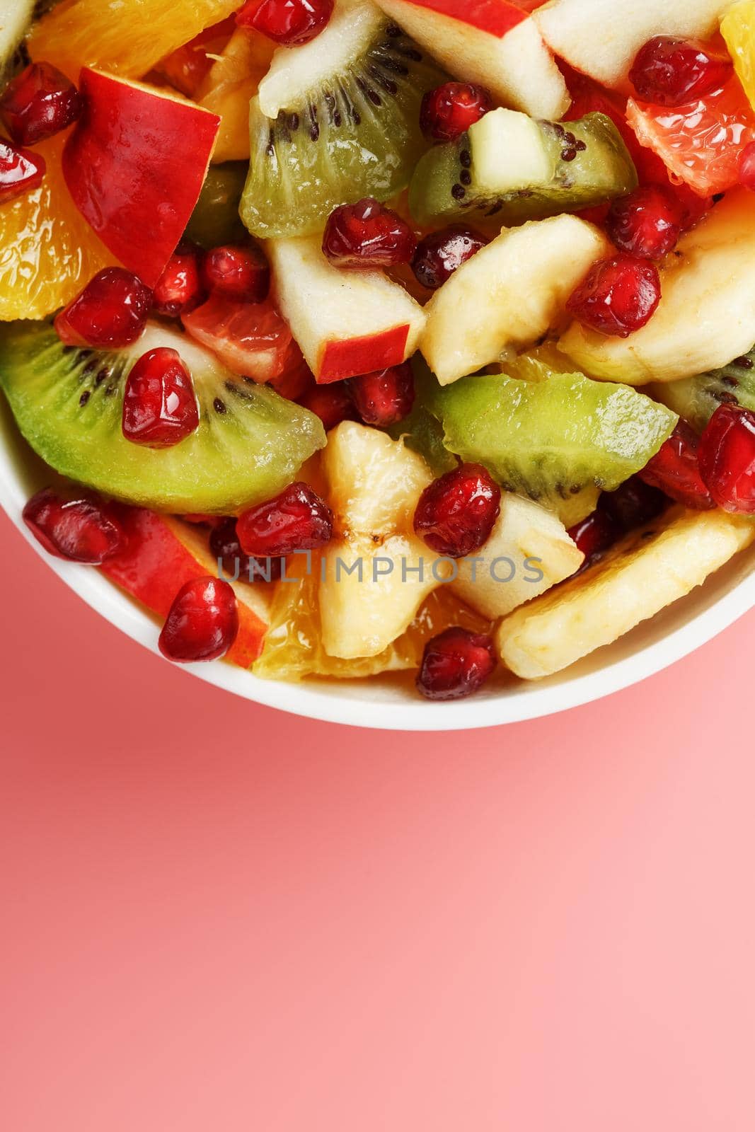 Salad of different juicy ripe fruits in a white cup on a pink background. Free space