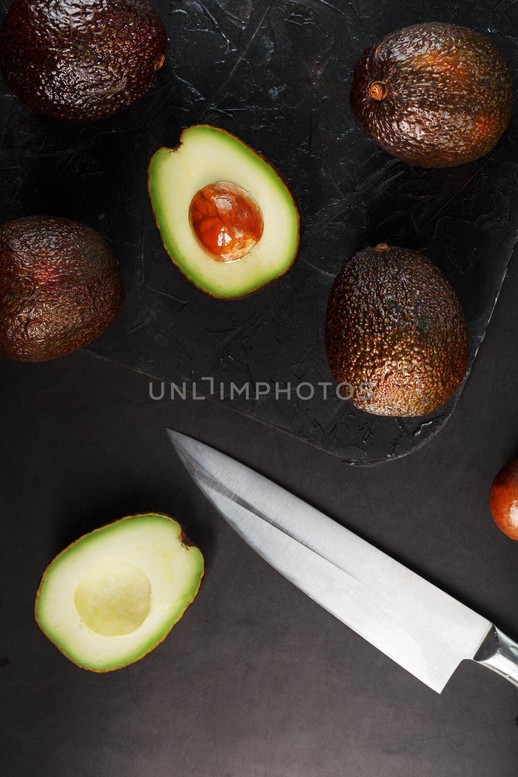 Whole and sliced avocados with a knife on a black textured black table, flat masonry. by AlexGrec