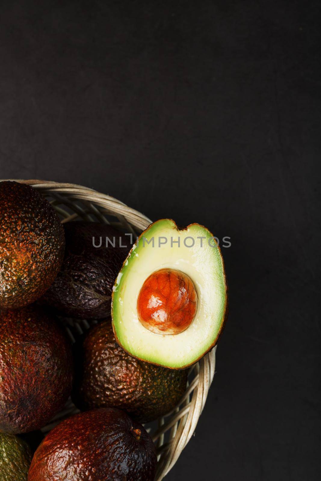 Ripe Hass avocado and a pitted slice in a basket on a black textured background. by AlexGrec