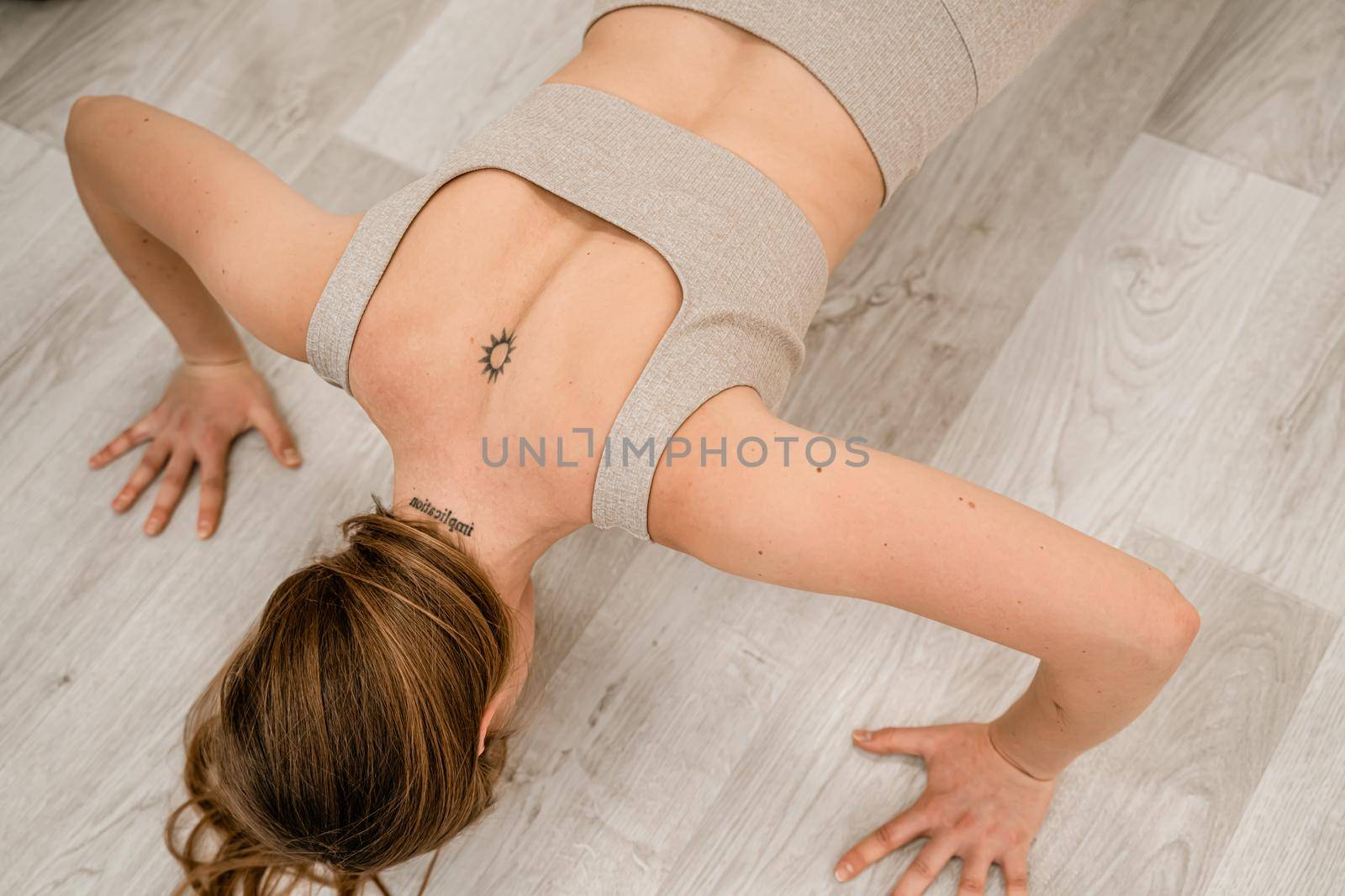 Top view of a muscular woman during push-ups. She is wearing a beige top and leggings, with a round tattoo between her shoulder blades. The concept of a healthy lifestyle. by Matiunina
