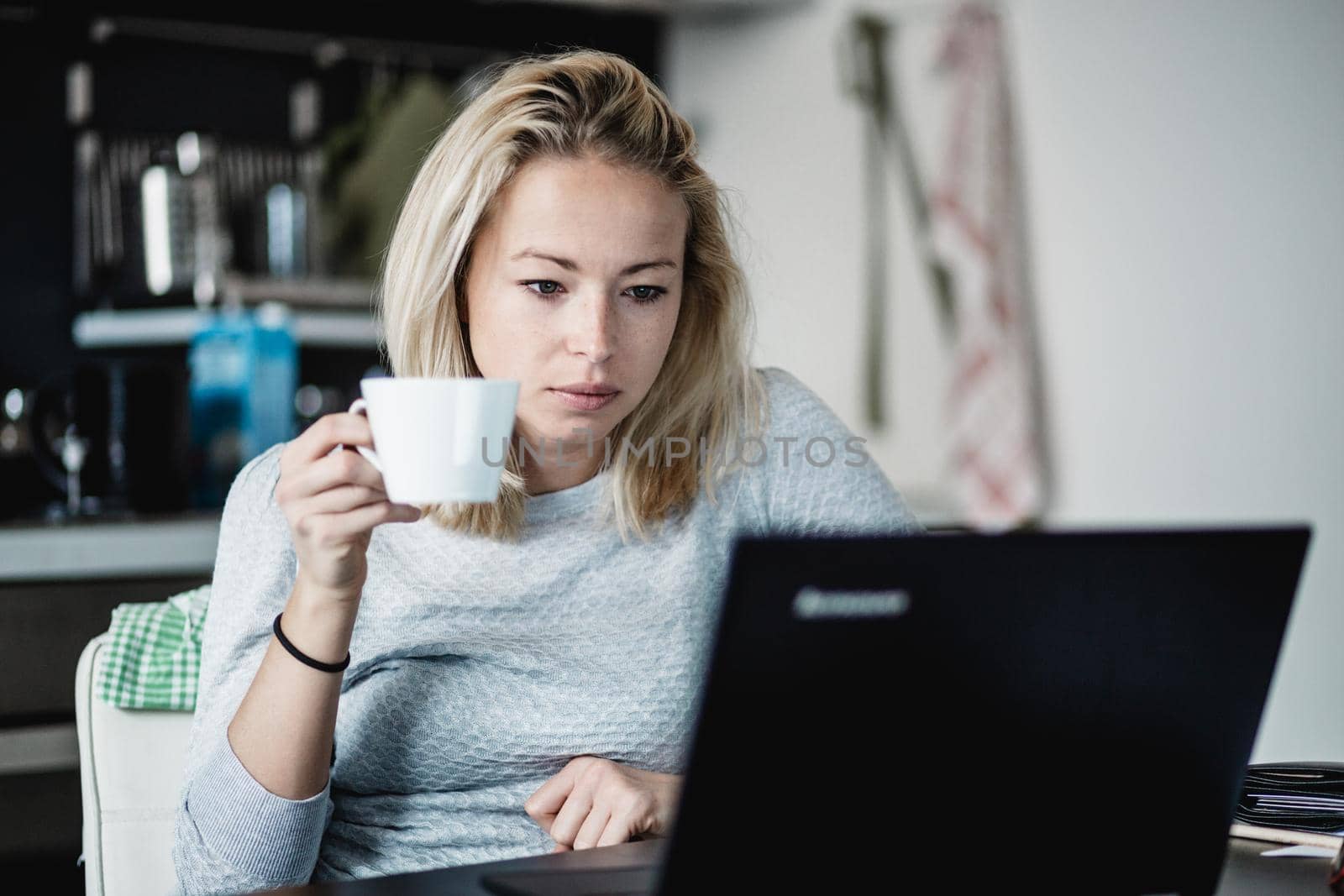 Female freelancer in her casual home clothing working remotly from her dining table in the morning. Home kitchen in the background. by kasto