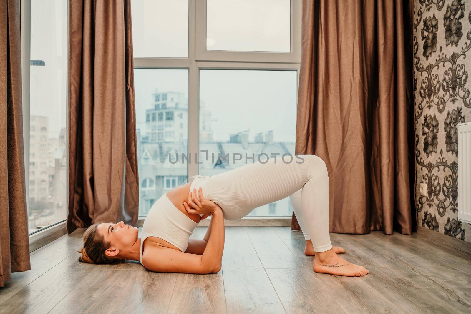 Young athletic attractive woman practicing yoga. Works out at home or in a yoga studio, sportswear, white pants and a full-length top indoors. Healthy lifestyle concept by Matiunina