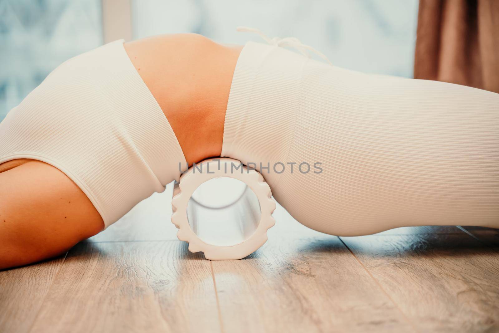 Adult athletic woman, in white bodysuit, performing fascia exercises on the floor - caucasian woman using a massage foam roller - a tool to relieve tension in the back and relieve muscle pain - the concept of physiotherapy and stretching training.