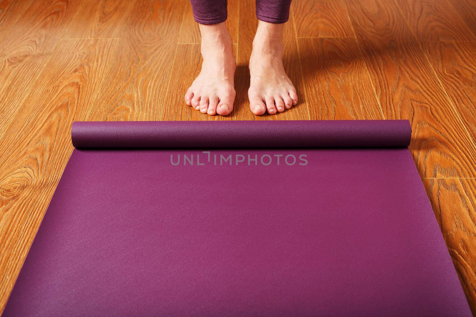 The girl's feet stand in front of an unfolded Yoga mat on the wooden floor. by AlexGrec